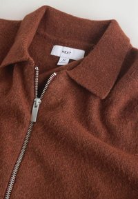 Veste en laine marron zippée avec col, taille moyenne, étiquette portant la mention "NEXT" et fermeture éclair métallique argentée partiellement ouverte.