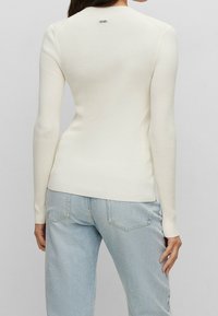 Ribbad, lätt creamfärgad tröja med långa ärmar och rund halsringning, med en liten logotyp nära halsen. Kombinerad med ljusblå jeans.