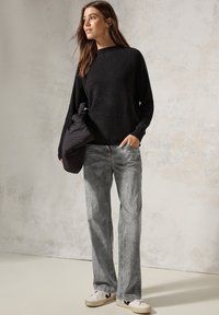 Zwarte textuursweater, lichtgrijze denim jeans met stippenpatroon, en witte sneakers met zwarte accenten; model staat tegen een neutrale achtergrond.