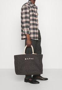 Borsa a tracolla nera intrecciata con manici beige e logo "MARNI". Il modello indossa una camicia a quadri bianca, nera e rossa, abbinata a pantaloni grigi.