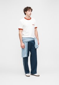 Λευκό μπλουζάκι με κόκκινο τελείωμα και λογότυπο, ανοιχτό μπλε πουλόβερ δεμένο γύρω από τη μέση, σκούρο μπλε παντελόνι με ρίγες στο πλάι και λευκά αθλητικά παπούτσια.