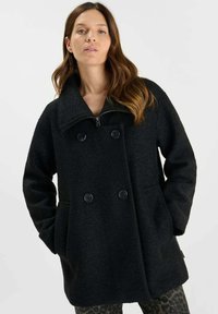 Zwarte wollen jas met een dubbelzijdig ontwerp, grote kraag, ritsluiting en twee voorzakken. Zachte textuur en een oversized pasvorm.