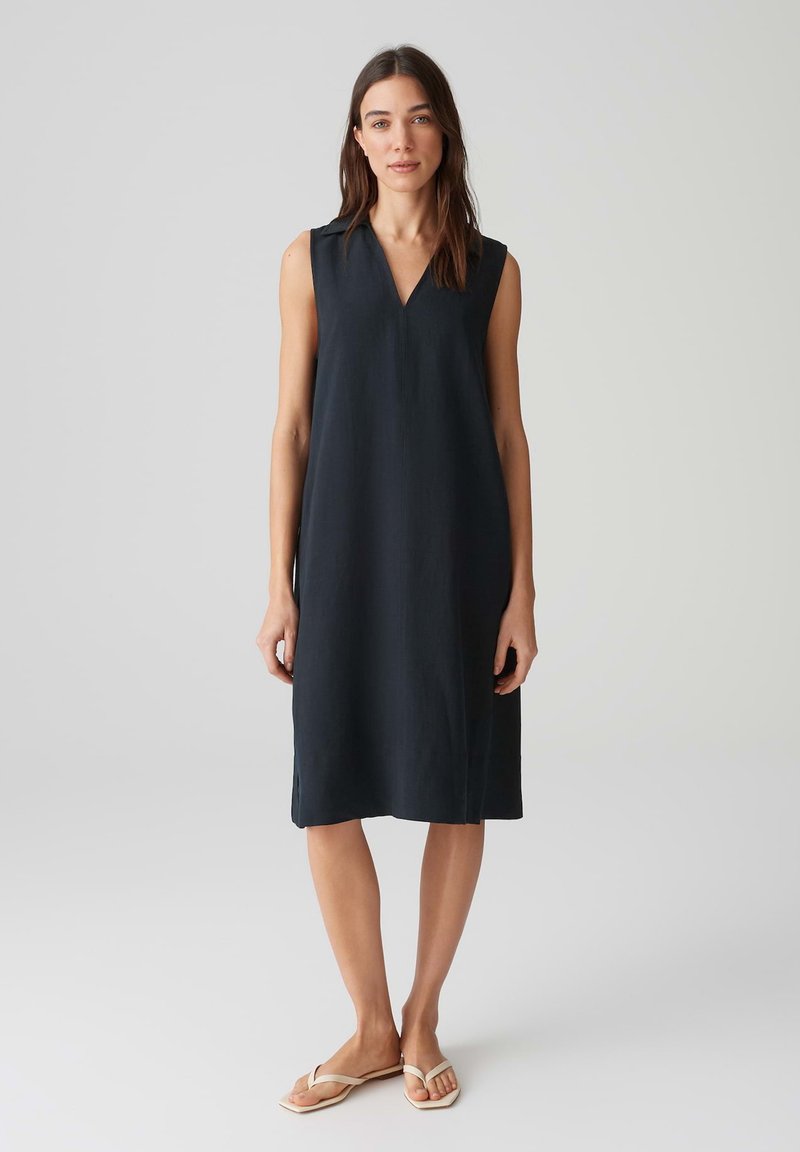 Sleeveless, navy dress with a V-neckline, knee-length design, made from a smooth fabric. Features side slits and minimal detailing.
