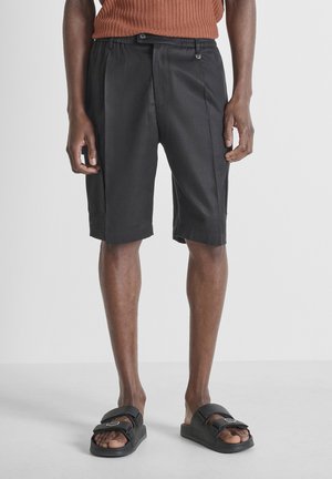 Hombre con pantalones cortos negros hasta la rodilla, camiseta sin mangas acanalada de color óxido y sandalias negras abiertas con hebilla, de pie sobre un suelo gris.