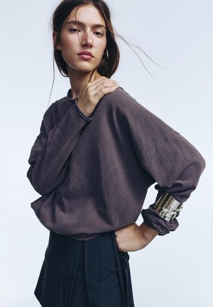 Ung kvinde med løst brunt hår iført en løs, mørk mauve langærmet top og sorte bukser, poserer med den ene hånd på skulderen og en bred sølvmanchet.