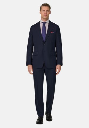 Mann trägt einen marineblauen Anzug mit zwei Knöpfen, gemusterte Krawatte, hell gestreiftes Hemd, Einstecktuch und schwarze Lederschuhe, steht frontal zur Kamera.