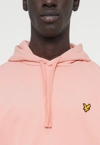 Ανοιχτόροζ φούτερ με κουκούλα από μαλακό ύφασμα, με τσέπη καγκουρό και κίτρινο κεντημένο λογότυπο στο στήθος, καθώς και ρυθμιζόμενα κορδόνια.