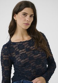 Vrouw met lang donker haar, draagt gouden ringoorbellen en een navyblauwe bloemenkant langemouwen top tegen een effen lichte achtergrond.