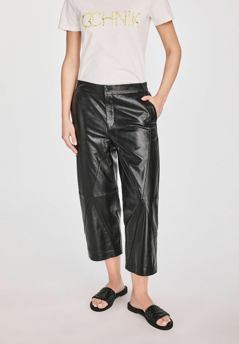 Czarne skórzane spodnie w kroju cropped o gładkiej fakturze i ukośnych szwach. W zestawie z białym t-shirtem i czarnymi plecionymi klapkami.