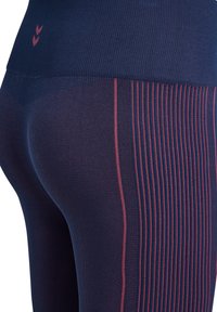 Zbliżenie granatowych legginsów z pionowymi czerwonymi paskami i logo w kształcie szewrona na górnej części biodra.