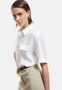 White short-sleeve shirt with a collar, featuring two front pockets. Worn with beige high-waisted pants and a black belt, smooth fabric finish.