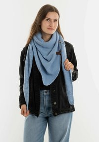 Lichtblauwe gebreide sjaal met een gestructureerd patroon, gedrapeerd in een driehoekige vorm, met een klein bruin label. Model draagt een zwarte jas.