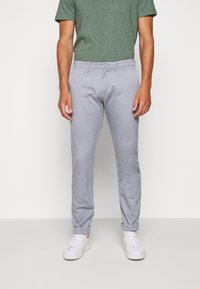 Světlé šedé kalhoty s úzkým střihem, ohrnutými lemy, předními kapsami a zapínáním na jeden knoflík, doplněné zeleným tričkem s pruhy.