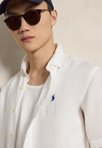 Mladý muž nosiaci slnečné okuliare s opáleným rámom, námornícku čiapku, bielu košeľu s otvoreným golierom a modré polo logo, a biele tričko pod ňou na neutrálnej pozadí.