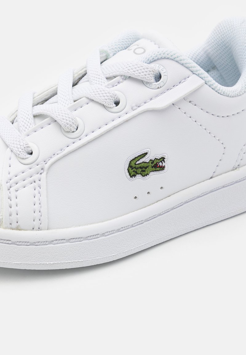 Sneaker en cuir blanc avec bout rond, lacets plats blancs, logo crocodile brodé vert et perforations minimales sur les côtés.