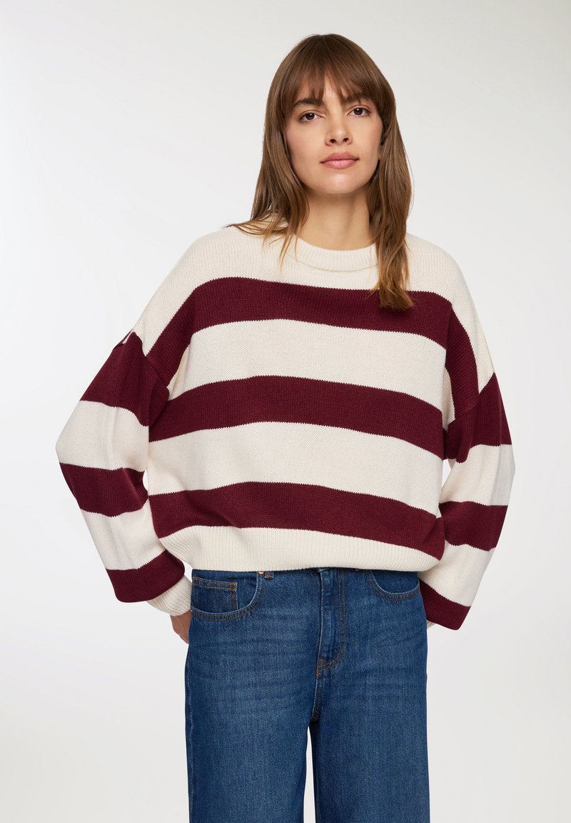 Sweter w burgundowe i kremowe paski, z opadającymi ramionami, okrągłym dekoltem i prążkowanymi mankietami, zestawiony z niebieskimi dżinsami o wysokim stanie.