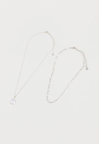 Deux colliers en argent sur fond blanc, l'un avec un pendentif en cristal en forme de goutte, l'autre avec des maillons ovales en cristal.