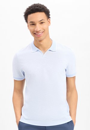 Lachende jonge man met kort krullend haar, gekleed in een lichtblauw polo met korte mouwen en donkerblauwe broek, tegen een witte achtergrond.