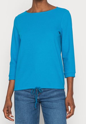 Vrouw draagt een felblauw topje met lange mouwen, een ronde hals en een tunnelkoord aan de zoom, gecombineerd met blauwe jeans.
