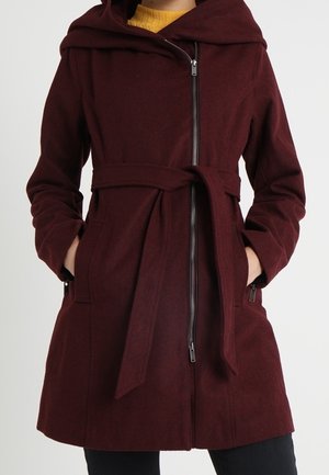 Vrouw draagt een bordeauxrode jas met ceintuur, diagonale rits en kap, handen in de zakken, over een mosterdgele top.