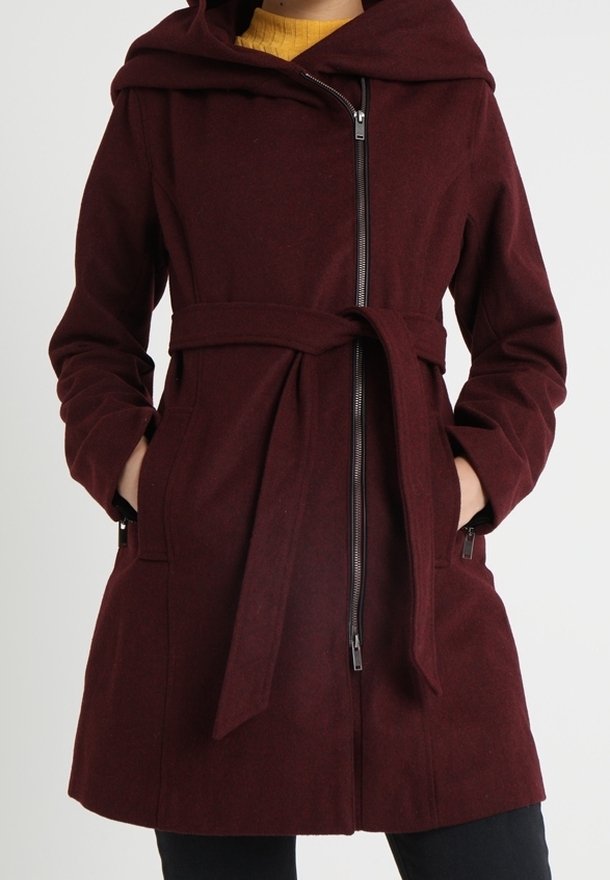 Femme portant un manteau bordeaux ceinturé avec une fermeture éclair diagonale et une capuche, mains dans les poches, sur un haut jaune moutarde.