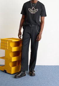 Camiseta negra con un logotipo gráfico colorido, pantalones negros y zapatos negros con adornos decorativos. Unidad de almacenamiento amarilla en el fondo.