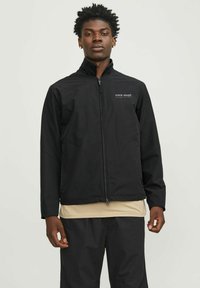 Zwarte ritsjack in lichtgewicht stof met een hoge kraag, voorzien van zijzakken en een logo op de linker borszijde. Gedragen over een beige shirt.