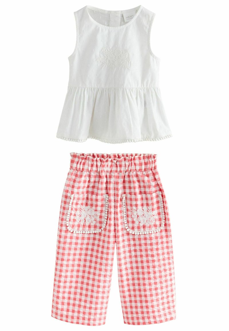 Biela bezrukávová blúzka so zberaným pásom a kvetinovou výšivkou; skombinovaná s červenými kockovanými nohavicami s vreckami lemovanými bielou čipkou.