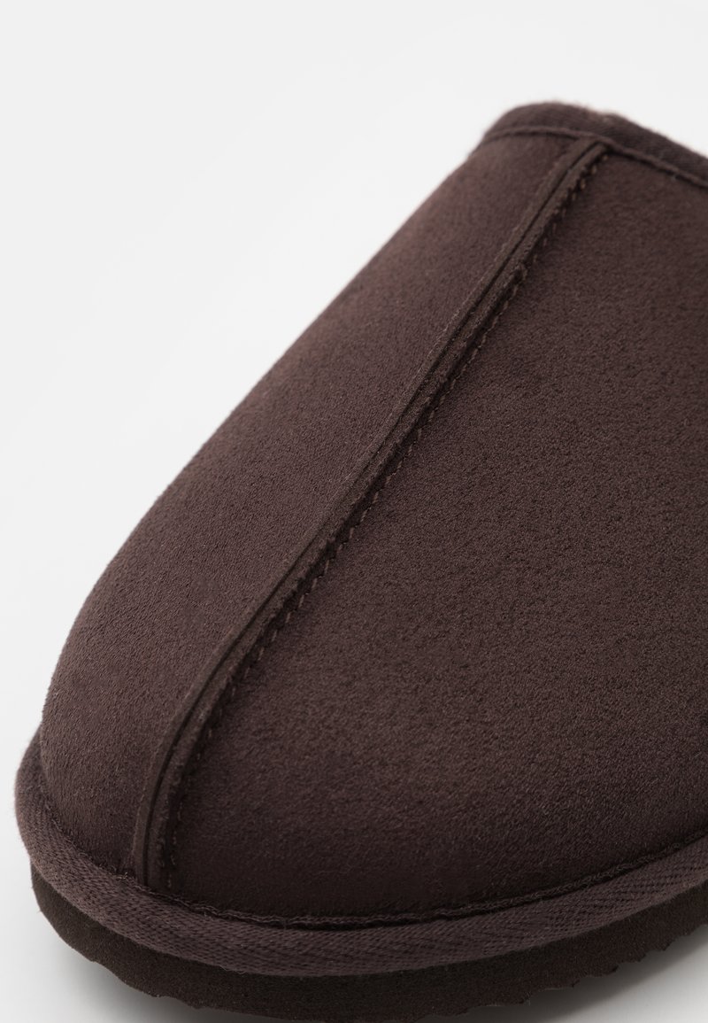 Zapatilla de ante marrón con diseño de punta redondeada, que presenta una costura prominente a lo largo del cuerpo y una superficie texturizada. Suela suave y acolchada.
