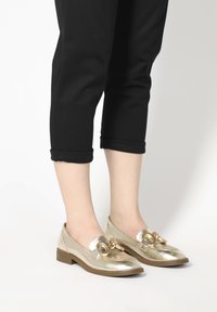 Zlaté metalické loafers s kulatým kovovým akcentem, plochou podrážkou a mírně špičatou špičkou. Jsou zkombinovány s ohrnutými černými kalhotami.