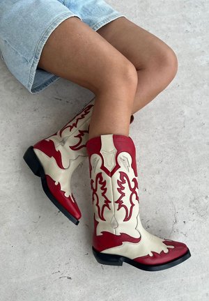 Bottes de cowboy rouges et crème avec des motifs enflammés, bouts pointus, tige de hauteur moyenne et semelles en caoutchouc noir résistant.