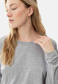 Grijze langmouwen shirt met een ronde hals, zachte textuur en geribbelde kraag, aangevuld met een hand die bij de hals rust en een grote oorbel.