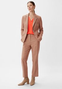 Terrakotta-karierter Anzug mit einer taillierten Blazerjacke und schmalen Hosen, kombiniert mit einem lebhaften orangefarbenen Top. Beige Knöchelstiefel runden den Look ab.