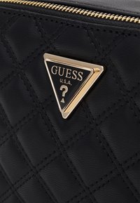 Sort quiltet lædertaske med en trekantet guld- og sort logo-plade. Lynlås med guld hardware. Tekstureret overflade og slankt design.