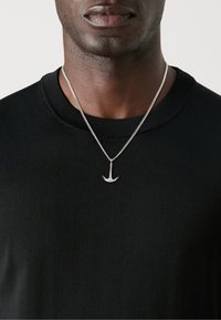 Chemise en maille noire avec un col ras du cou et un pendentif en forme d'ancre en argent sur une chaîne, présentant une finition lisse et des accents détaillés.