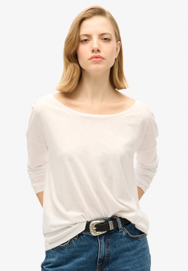 Witte blouse met lange mouwen en een brede halslijn, gecombineerd met hoge blauwe spijkerbroeken en een zwarte riem met een zilveren decoratieve gesp.