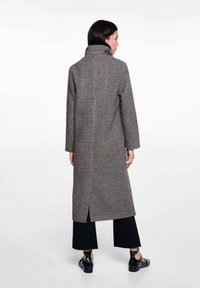 Houndstooth-patterned, gray long coat with a high collar, open back vent, and long sleeves, worn over black wide-leg pants and shoes.