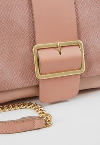 Sac à main rose texturé avec une lanière en cuir lisse, une boucle rectangulaire dorée et un détail chaîne dorée sur une surface blanche.