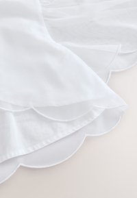 Tela blanca con un detalle de dobladillo en forma de ondas. El material es ligero y semitransparente, exhibiendo una textura suave.