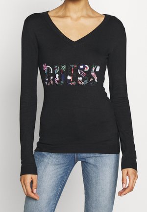 Kobieta ubrana w czarny, długi sweter z dekoltem w serek, ozdobiony kolorowym kwiatowym wzorem i cekinowym logo "GUESS", w połączeniu z jasnoniebieskimi dżinsami.