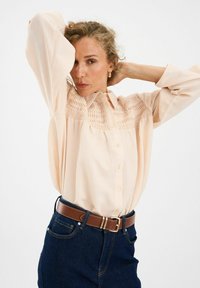 Kerge virsiku värvi nööbitav särk, mille kaelas on volditud detailid, koos tumedate teksade ja pruuni nahkvööga.