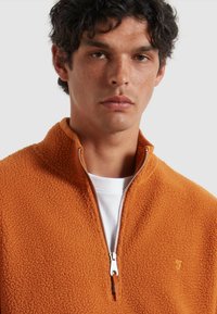 Pullover arancione in fleece con mezzo zip e superficie texturizzata, dotato di una cerniera argento e di un piccolo logo ricamato sul petto.