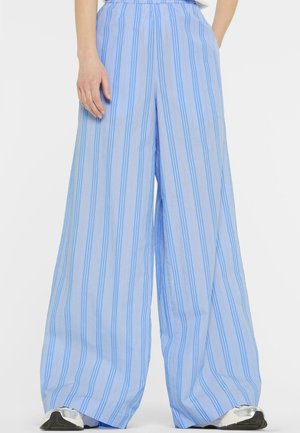 Woman wearing wide-leg, high-waist blue pants with vertical stripes and silver sneakers, standing with one hand in pocket.