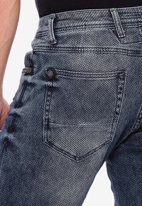 Jean en denim bleu présentant un motif texturé et délavé avec des coutures contrastées. Comprend des poches arrière doubles et des boutons en métal.