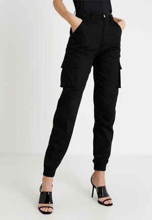 Femme portant un pantalon cargo noir avec des bords élastiques et des sandales noires à talons ouverts, debout les mains dans les poches.