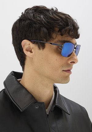 Uomo con capelli corti neri che indossa occhiali da sole blu riflettenti e una giacca di pelle nera, rivolto verso destra su uno sfondo uniforme.