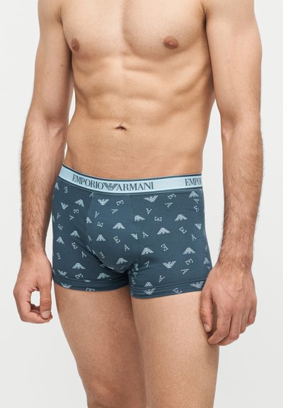 Αρσενικός κορμός που φορά μπλε boxer briefs Emporio Armani με μοτίβο λογότυπου σε όλη την επιφάνεια και branded ζώνη μέσης, στέκεται μπροστά σε απλό φόντο.