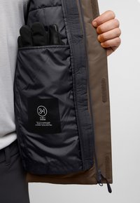 Geïsoleerde 3-in-1 jas met een donkergrijze binnenkant, bruine buitenkant, rits sluiting en zwarte handschoenen in een zak. Verzorgingslabel zichtbaar.