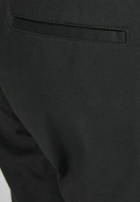 Zwarte broek met een close-up van de achterzak. De stof is glad en heeft een subtiele textuur met een gestikt naaigedeelte rond de zak.