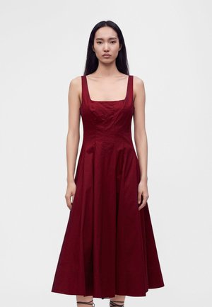 Femme aux longs cheveux foncés portant une robe midi sans manches rouge foncé avec un décolleté carré, debout devant un fond blanc uni.